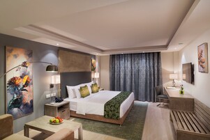 A hotel room with a large bed, a desk, a chair, a lamp, and artwork on the walls.