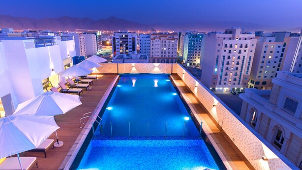 Una piscina en la azotea con sillones de descanso y sombrillas, rodeada de edificios altos.