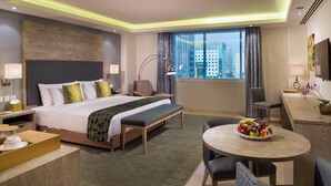 Habitación de hotel con una cama grande, un escritorio, una silla, una mesa redonda con fruta y vistas a edificios a través de la ventana.