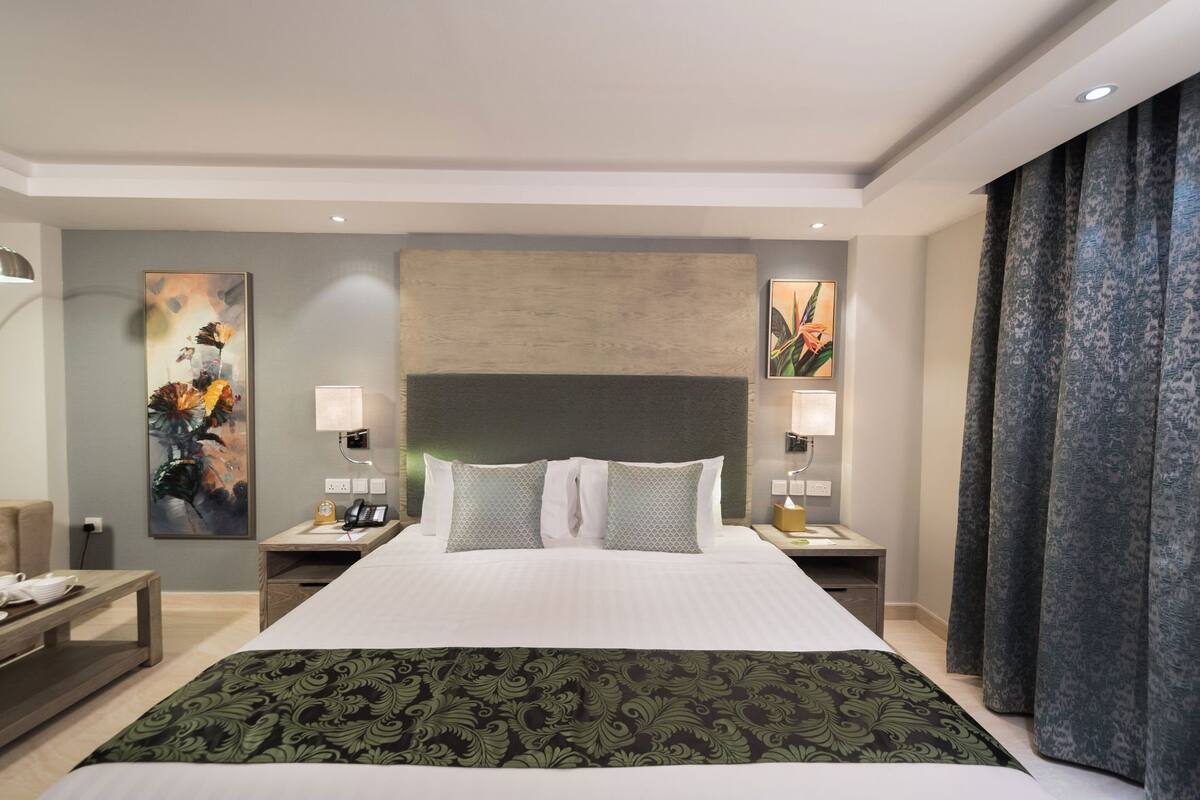 Una habitación de hotel con una cama grande, dos mesitas de noche, un buró, un cuadro en la pared y una ventana con cortinas.