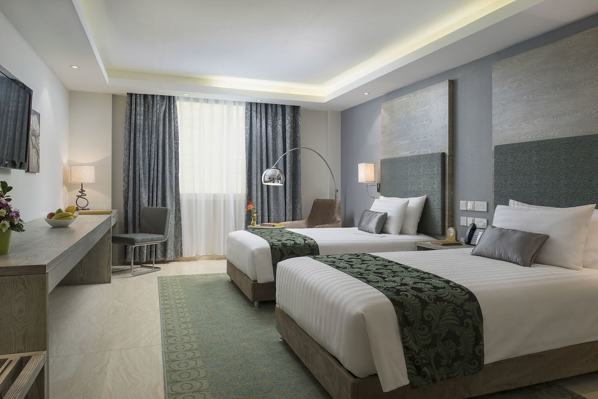 A hotel room with two beds, a desk, a chair, a lamp, and a view of the outside.