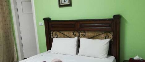 Un lit avec une tête de lit en bois, une literie blanche et une serviette violette en forme de cygne. Il y a deux tableaux encadrés au mur, une porte et une table de chevet avec une boîte de mouchoirs.