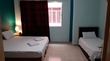 Una habitación con dos camas, una ventana con cortinas, una mesita de noche con lámpara y una puerta.