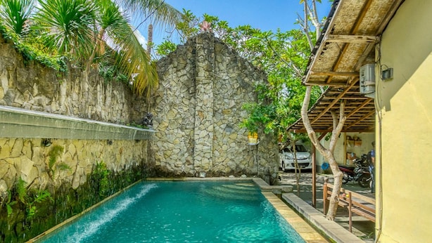 A swimming pool with clear blue water, surrounded by a stone wall and lush greenery. - Lagas Hostel (Ubud)