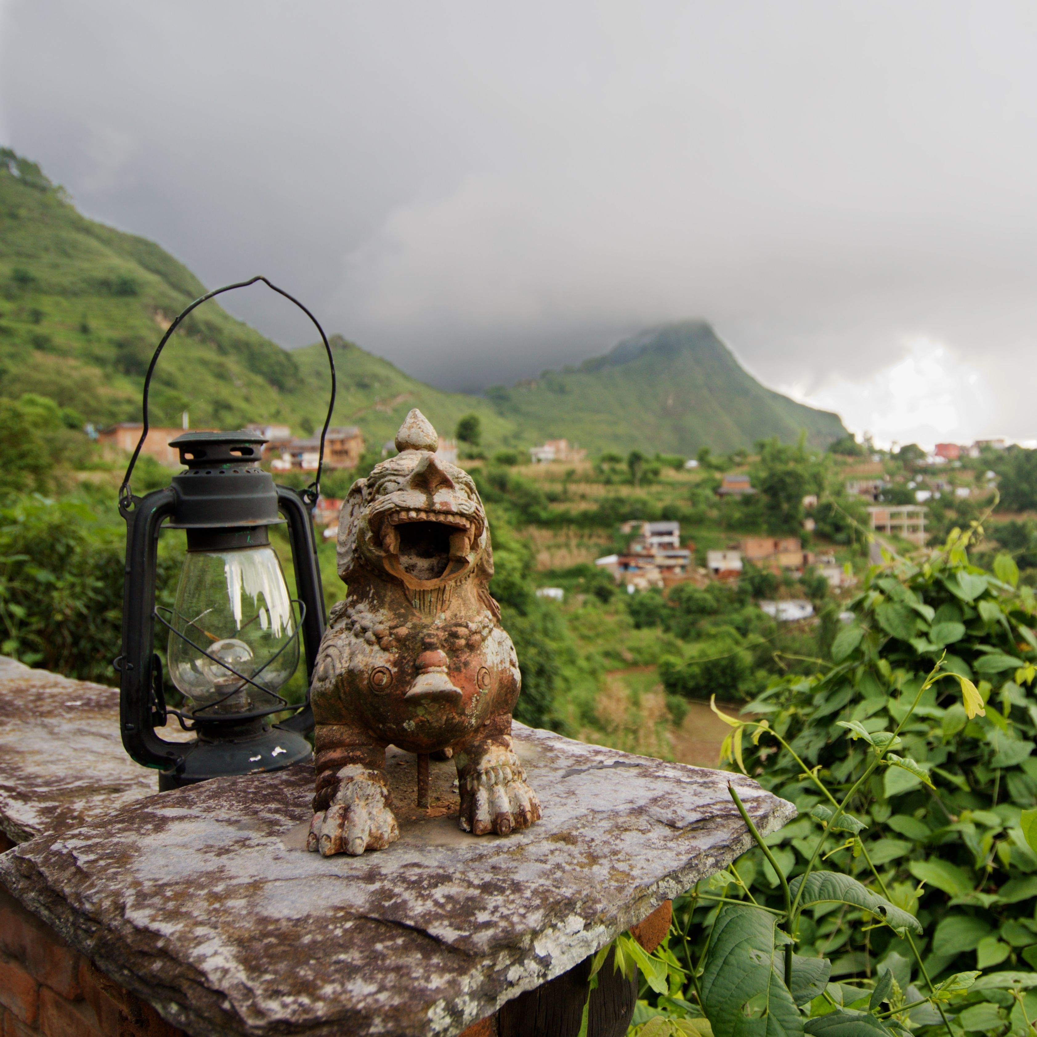 A stone lion statue with a lantern on a ledge, overlooking a village and mountains.