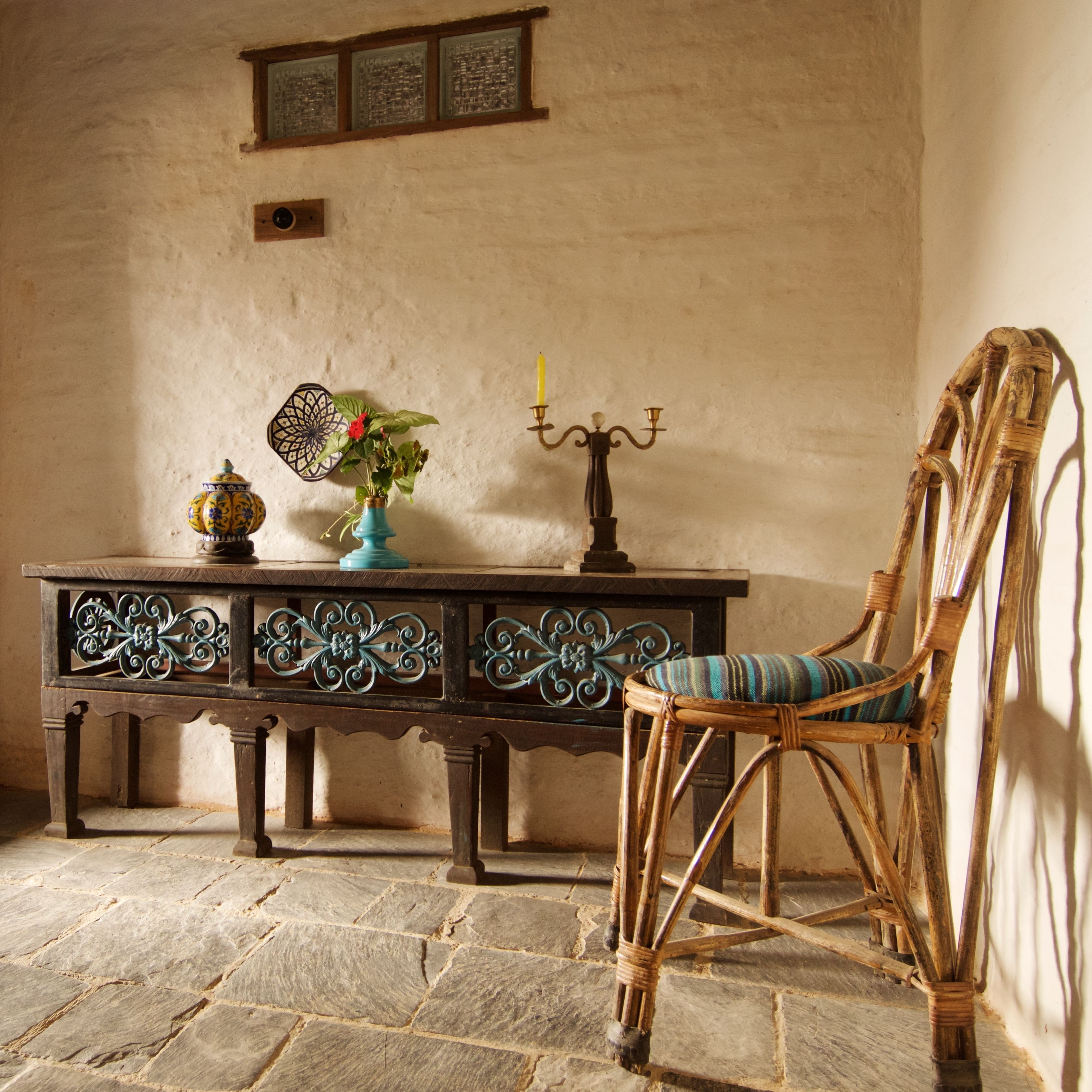 Una habitación rústica con suelo de piedra, una mesa de madera con intrincados grabados, un sillón de mimbre y un cuadro colgado en la pared.