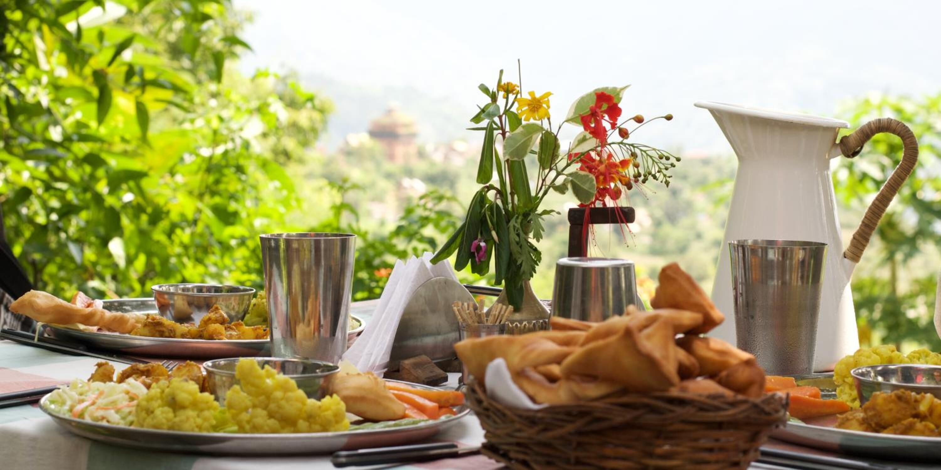 A table set with various dishes, including fried items, vegetables, and a pitcher, with a view of greenery and a distant structure.