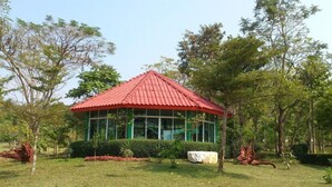 Een paviljoen met een rood dak en grote ramen, omgeven door een gazon en bomen.