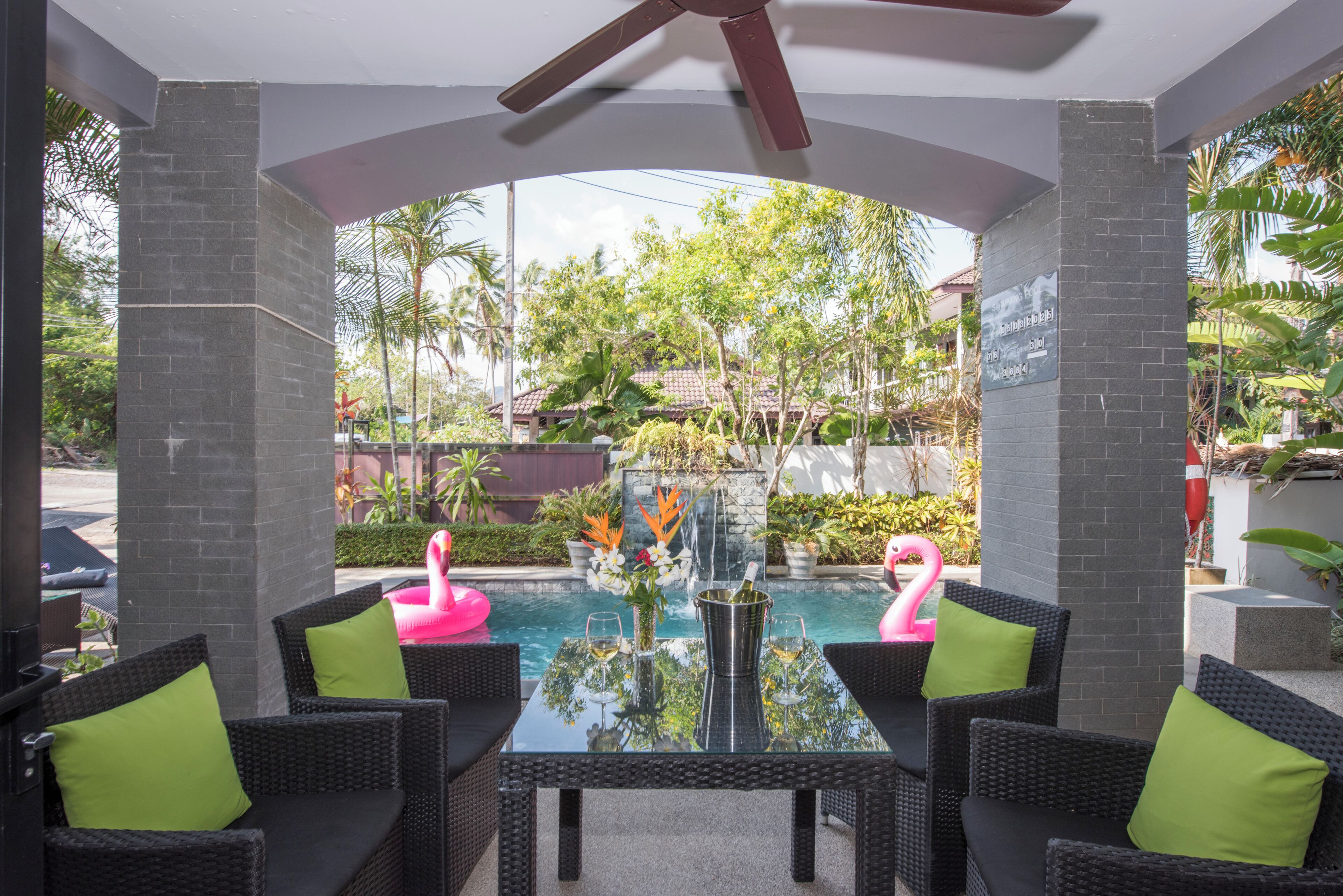 An outdoor seating area with wicker furniture and a glass table, overlooking a pool with flamingos and tropical plants.