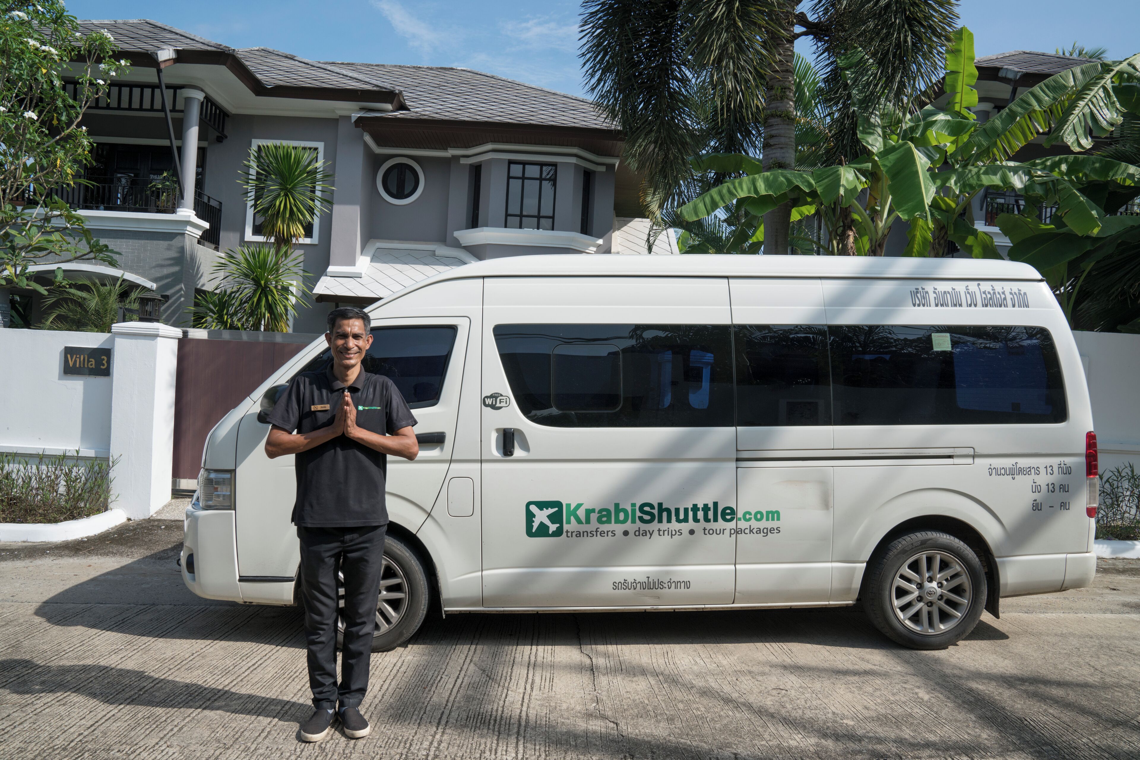 A man standing next to a white van with 'KrabiShuttle.com' branding, parked in front of a house with a gated entrance and tropical vegetation.