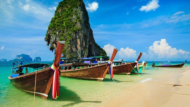 A beach with wooden boats tied to the shore, a large rock formation, and clear turquoise water.