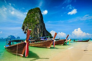 Une plage où des bateaux en bois sont amarrés au bord, une grande formation rocheuse et une eau turquoise cristalline.