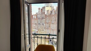 A room with a yellow armchair, a glass table, and a view of a cityscape through a large window with black curtains. - Hotel Heye 130 (Amsterdam)