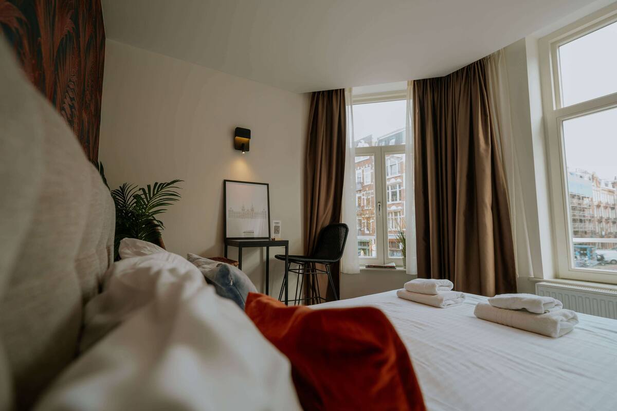 A hotel room with a bed, a desk, a chair, a potted plant, and a window with curtains.