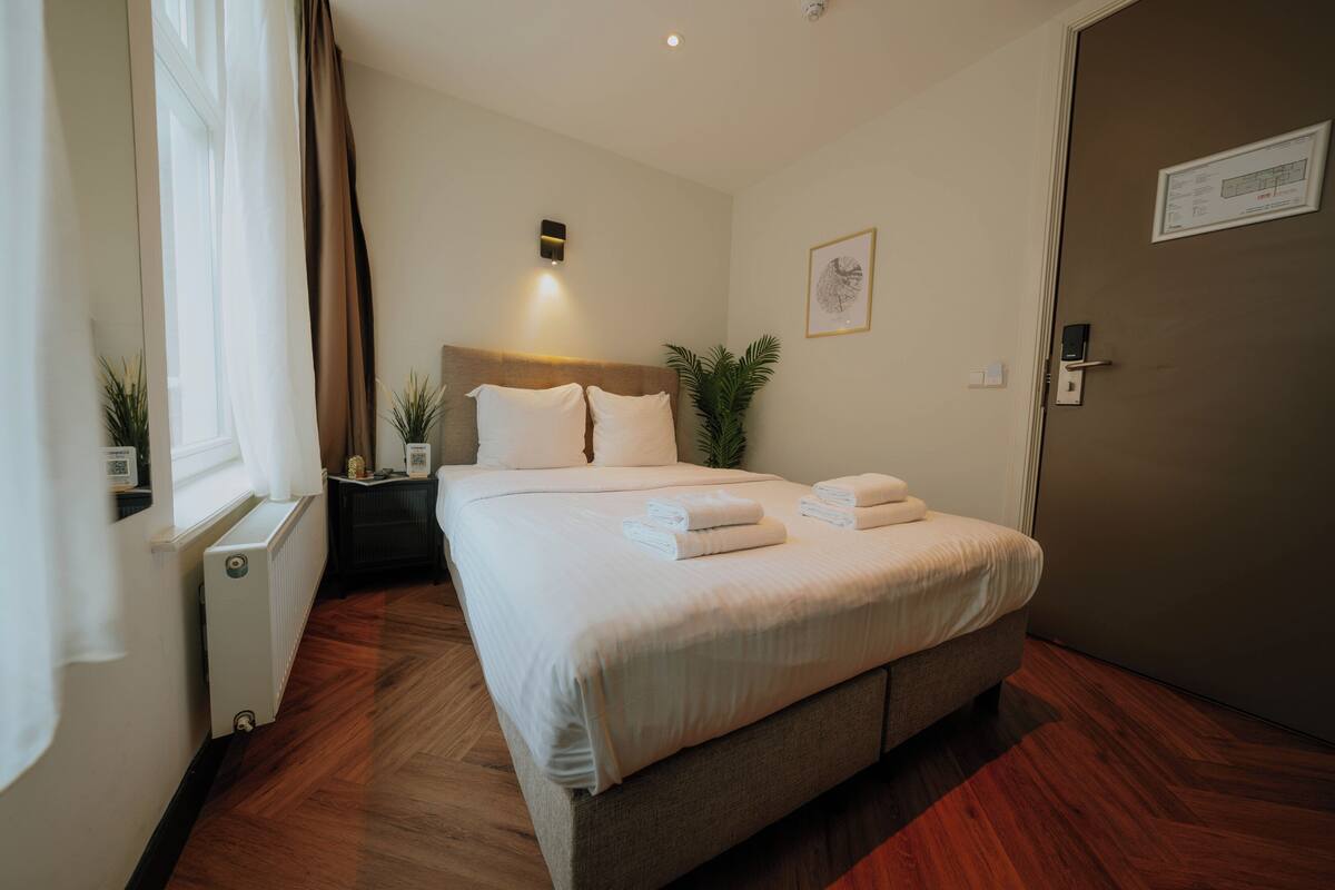 A neatly made bed with white linens and towels, a bedside table with a lamp, a radiator, and a door with a lock and a sign.