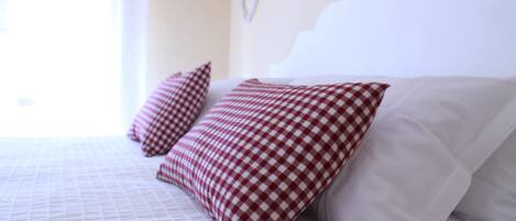 A neatly made bed with a checkered pillow and a plain pillow, a window with sheer curtains, and a wall-mounted light fixture.