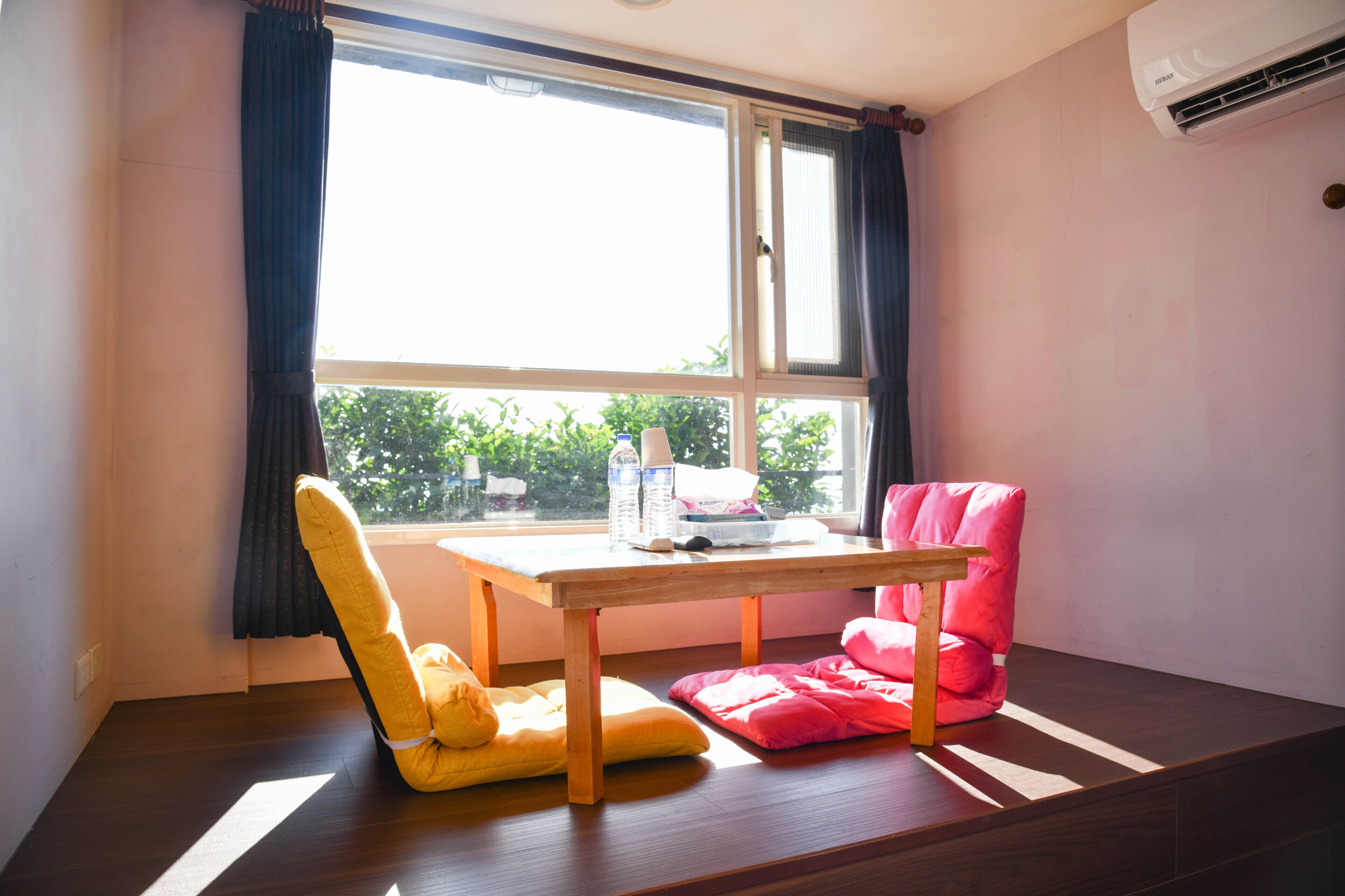 Une pièce avec une table en bois, une chaise jaune et une chaise rose. Il y a une fenêtre avec des rideaux et un appareil de climatisation fixé au mur.