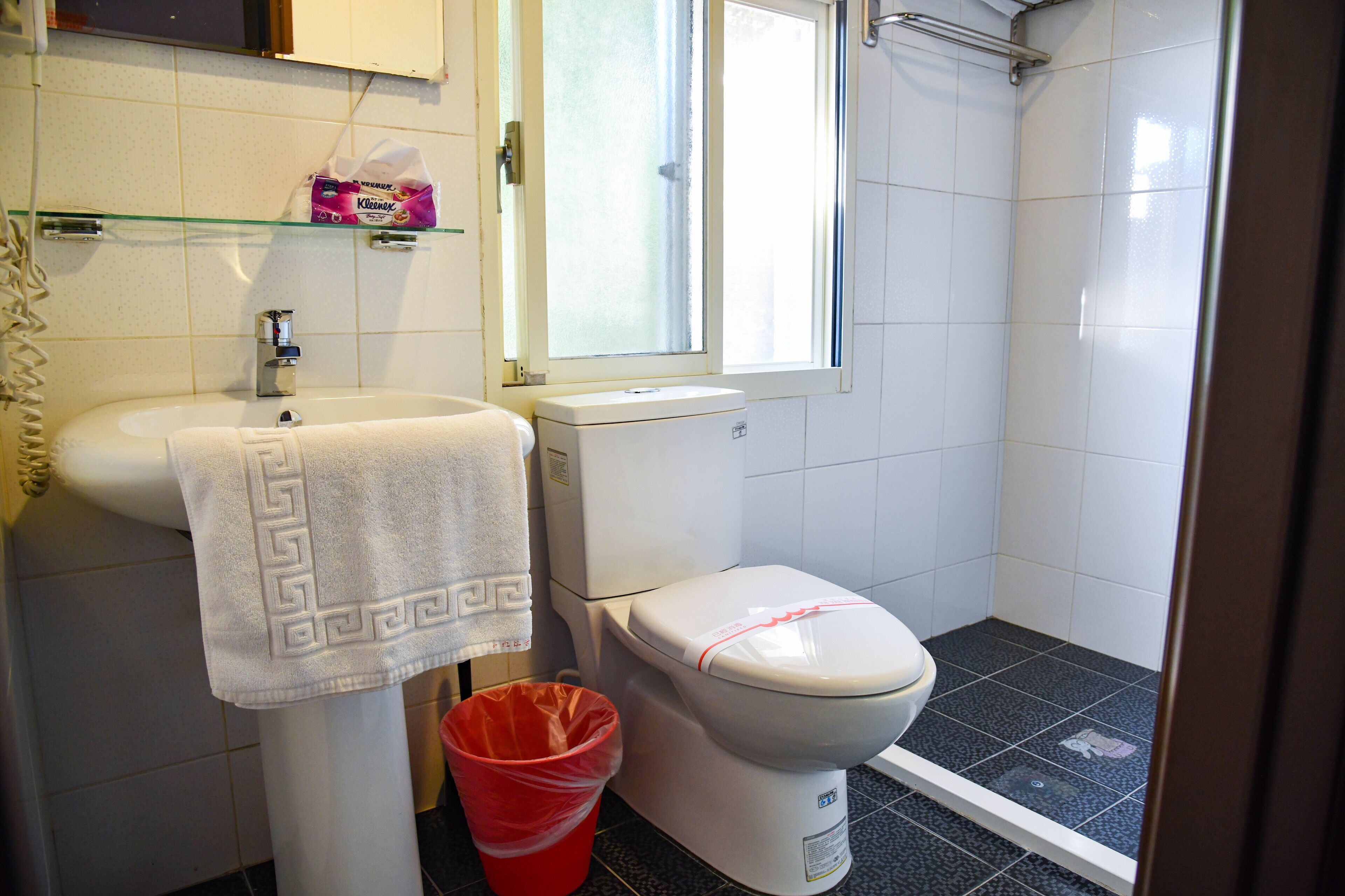 A bathroom with a white sink, toilet, and shower. There is a glass shelf above the sink with a pink box and a white towel hanging on the wall.