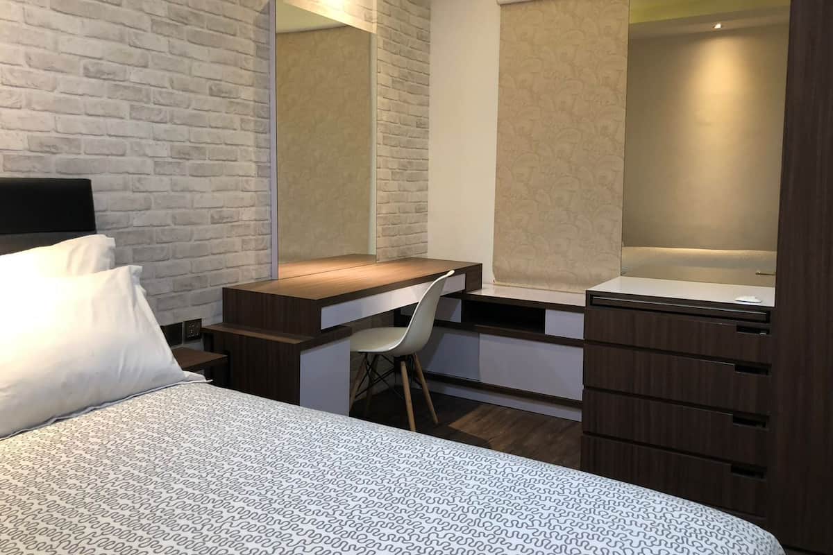 A modern hotel room with a brick-patterned wall, a wooden desk with a chair, a bed with a patterned bedspread, and a large mirror above the desk.