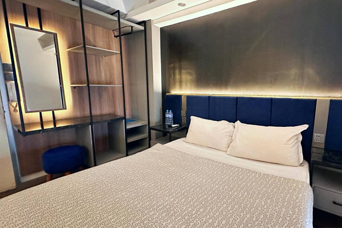 A hotel room with a large bed, a bedside table with a lamp, a small desk, a chair, and a wardrobe with a mirror.