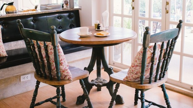 A dining area with a round table, two chairs, a bench, and a window with white shutters. - Royal Princess City (Lamphun)