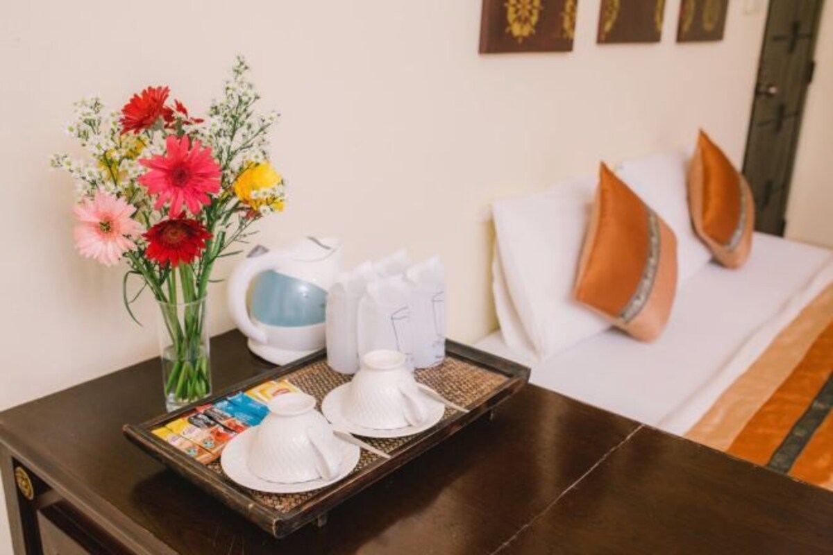 Habitación de hotel impecablemente ordenada, con una cama, almohadas, una bandeja con teteras blancas, un jarrón con flores coloridas y una mesita con una revista.
