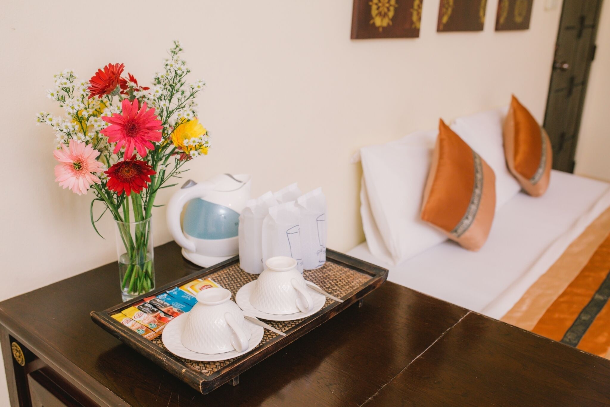 A hotel room with a bed, pillows, a tray with teapots, cups, and a vase of flowers.