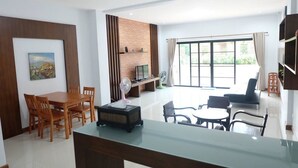 A modern living room with a dining area, a brick accent wall, a large window, and a TV. - Baan Hauen Inn (Chiang Mai)
