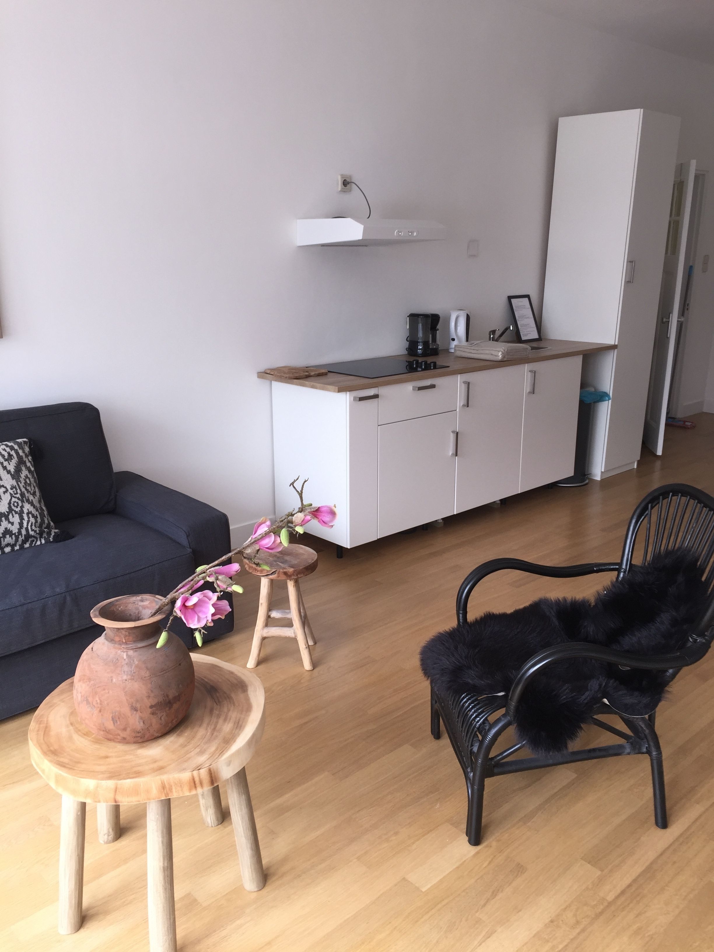 A modern living space with a sofa, a black chair, a small wooden table with a vase, and a kitchen area with cabinets and appliances.