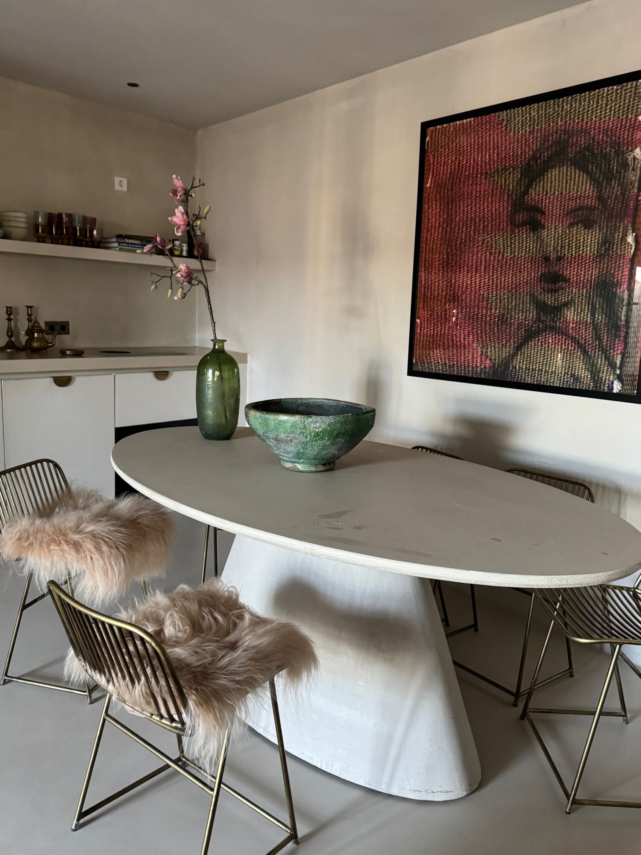 Een eetgedeelte met een ronde tafel, stoelen, een vaas met bloemen en een groot abstract schilderij aan de muur.