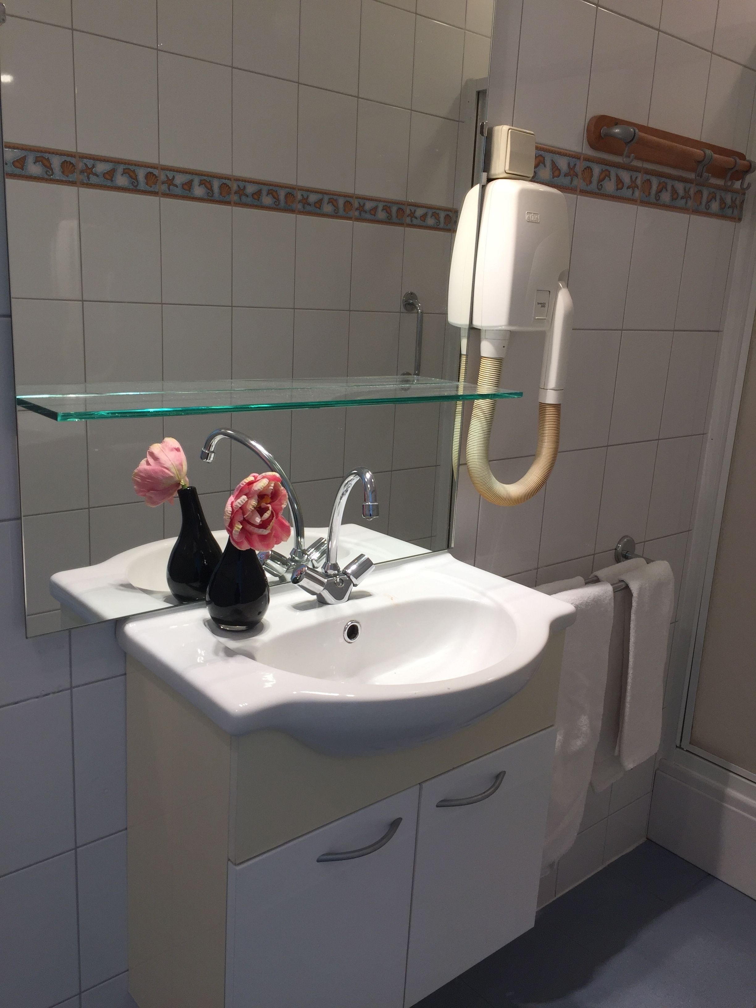 Un baño con lavamanos blanco, estante de vidrio, espejo y toallero.