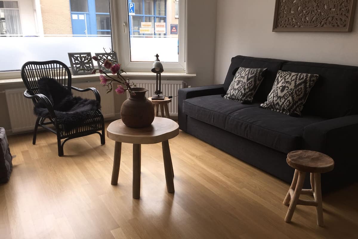 Un salon avec un canapé gris foncé, un fauteuil noir, une petite table ronde ornée d’un vase et un tabouret.