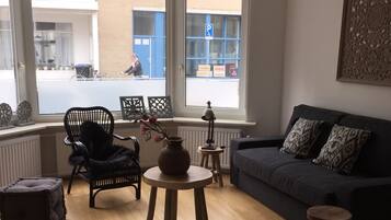 Un salon comprenant un canapé, un fauteuil, une petite table et une fenêtre donnant sur la rue.