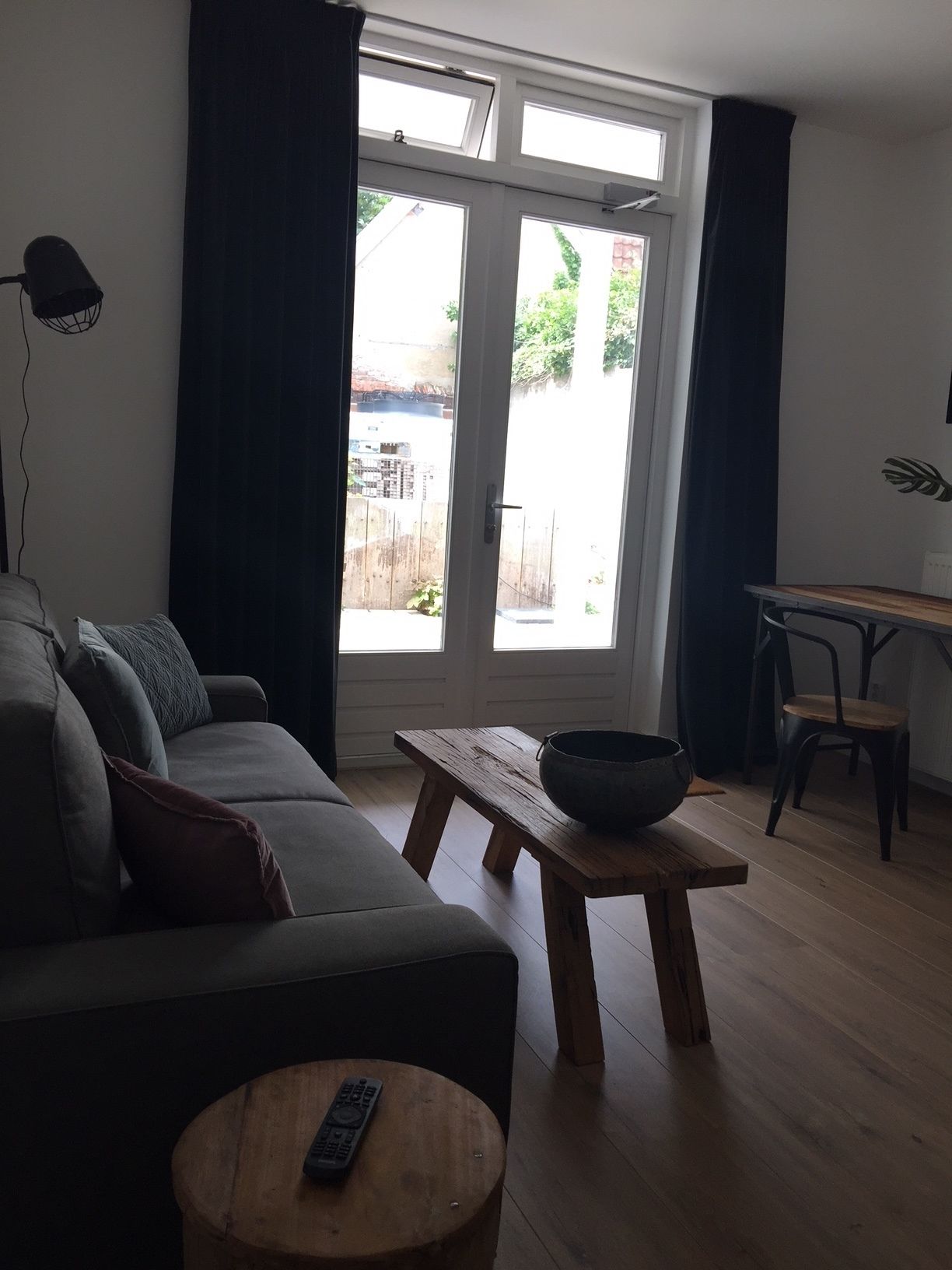Un salon comprenant un canapé, une table en bois avec un bol, une petite table ronde en bois avec une télécommande, et un coin repas avec des chaises et une table.