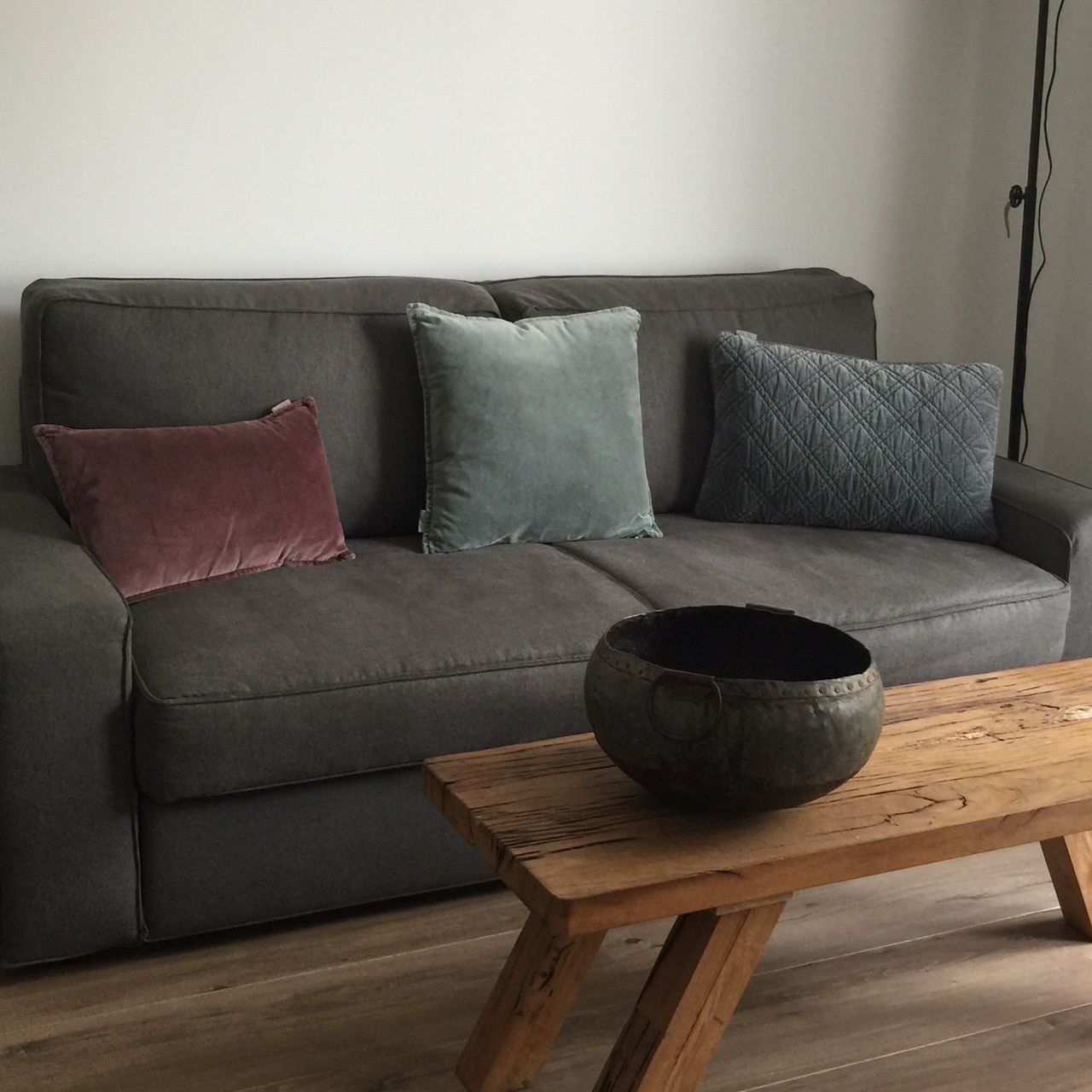 Un sofá gris con tres cojines de distintos colores y una mesa de centro de madera rústica con un cuenco oscuro.