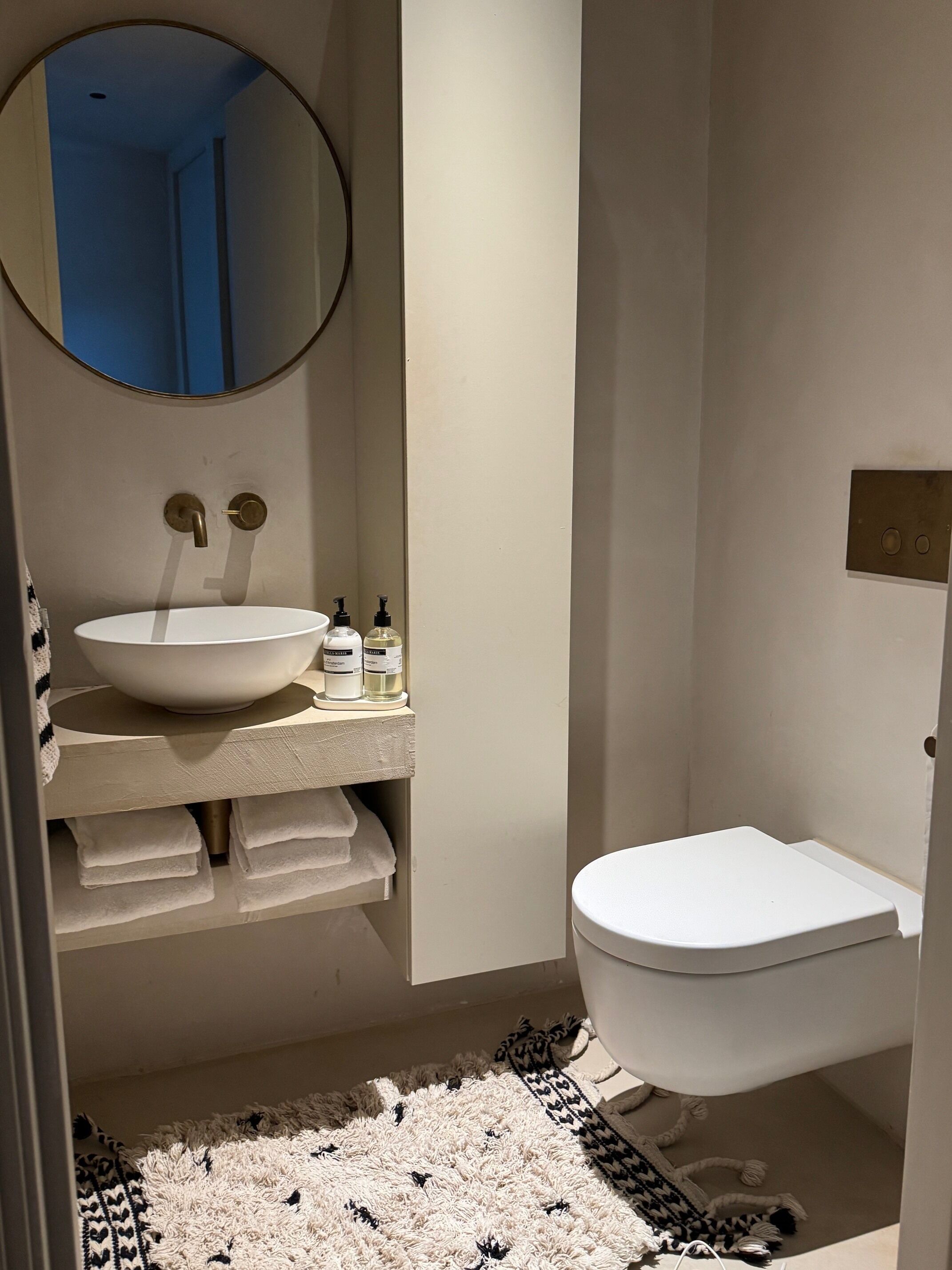 A bathroom with a white sink, a round mirror, a toilet, and a patterned rug.