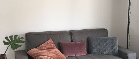 Un canapé gris avec trois coussins, une table d’appoint en bois avec un bol, une grande plante verte et un grand lampadaire.