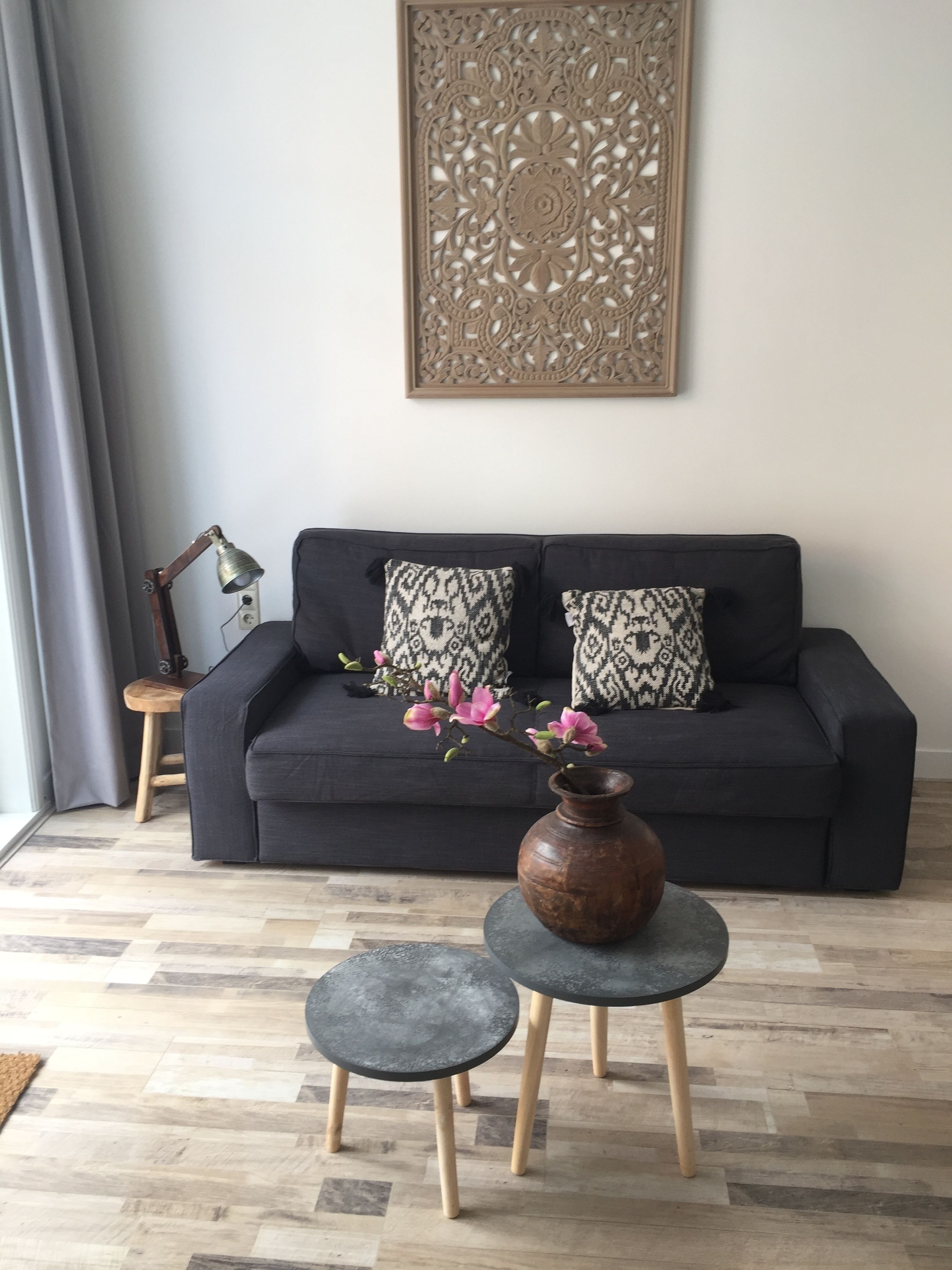 Un salon moderne avec un canapé foncé, des coussins à motifs, un élément décoratif mural, un vase avec des fleurs et deux petites tables.