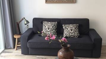Uma sala de estar moderna com um sofá escuro, almofadas estampadas, um objeto decorativo na parede, um vaso com flores e duas mesinhas.