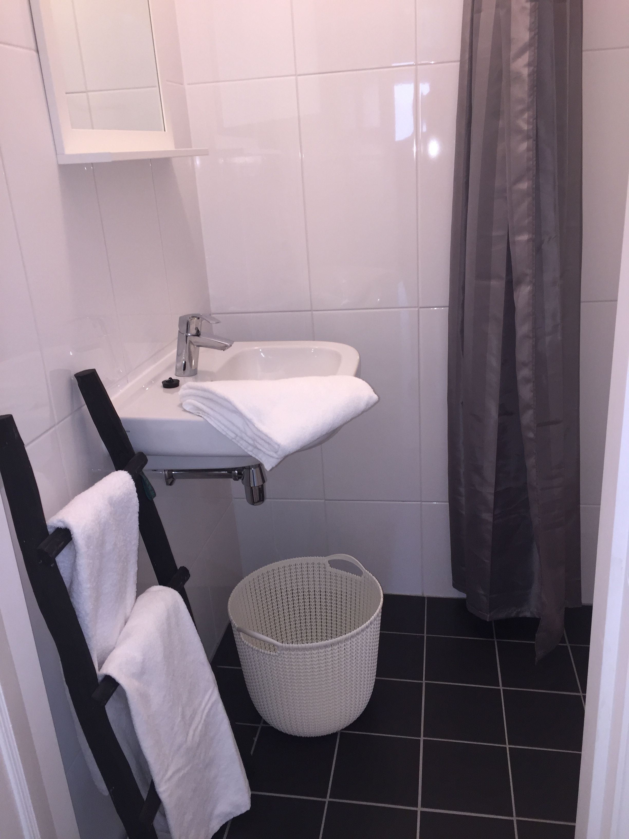 Un baño con lavamanos blanco, toallero negro, cesta blanca y cortina de ducha.