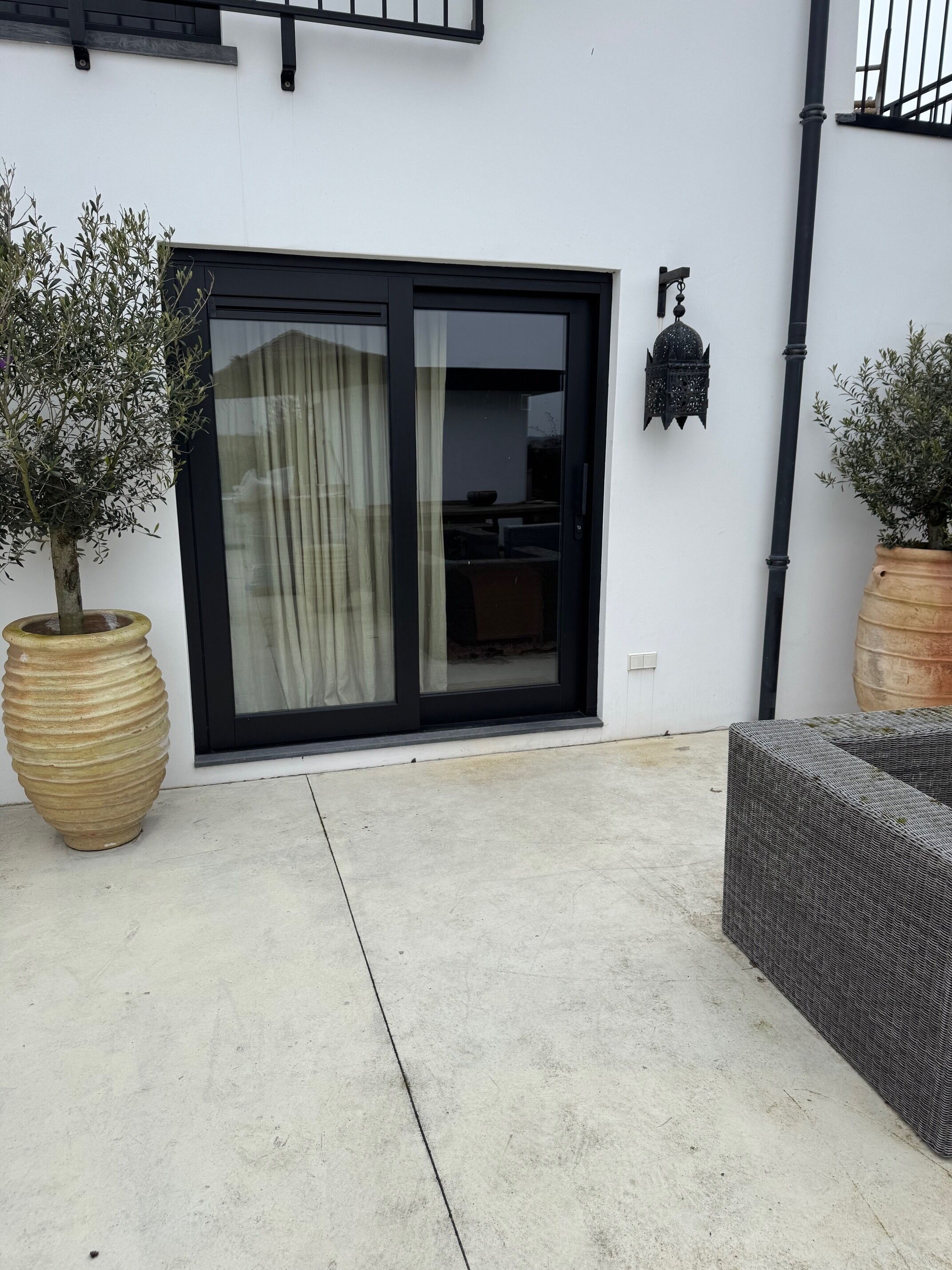 Un bâtiment moderne doté d’une grande porte-fenêtre coulissante, d’un olivier en pot et d’une lanterne décorative fixée au mur.