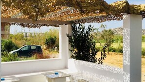 A covered outdoor seating area with white benches and a table, a car parked outside, and a clear sky. - Helios Beach Studios (Naxos)