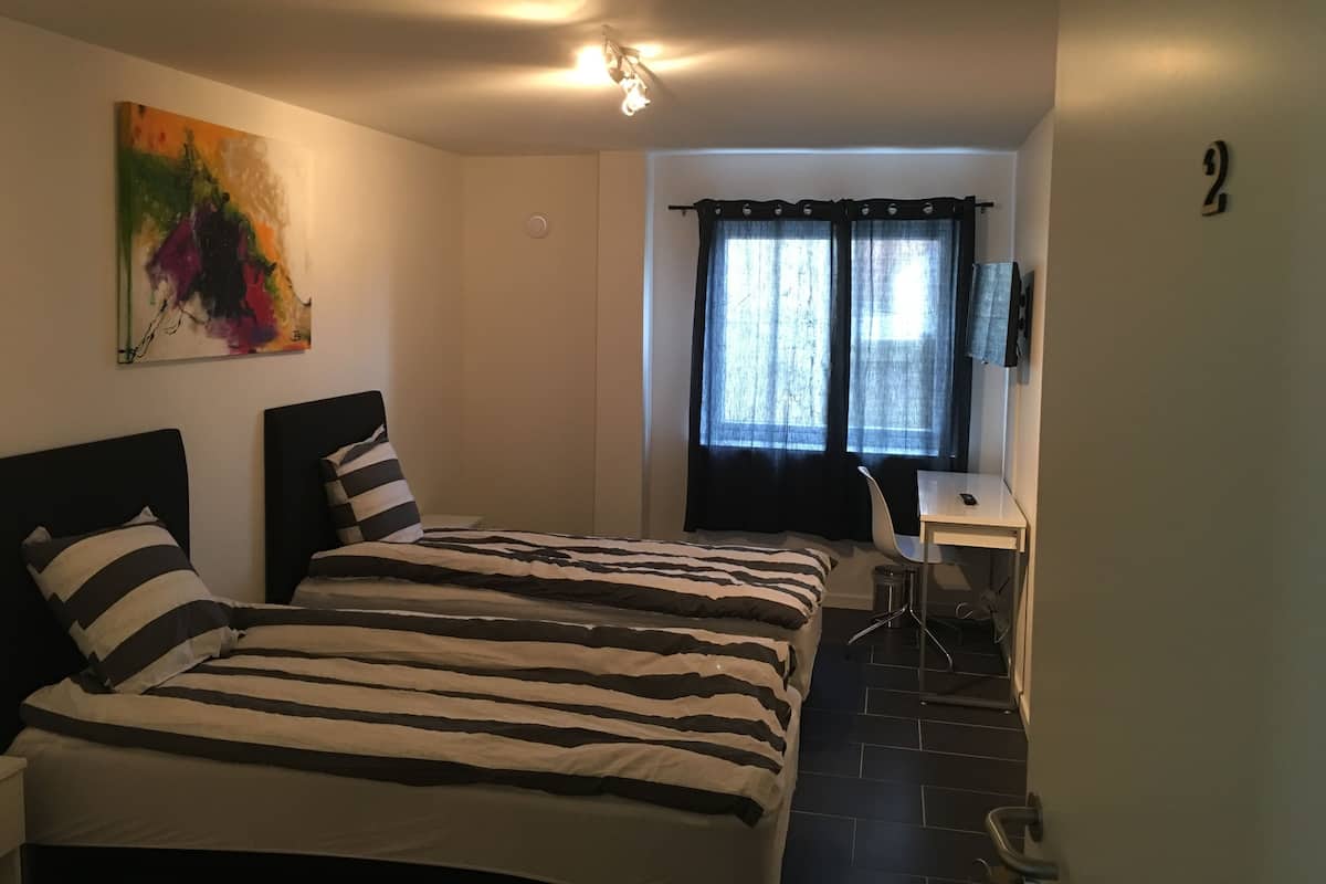 Una habitación con dos camas, un escritorio, una silla, un televisor y un ventanal con cortinas.