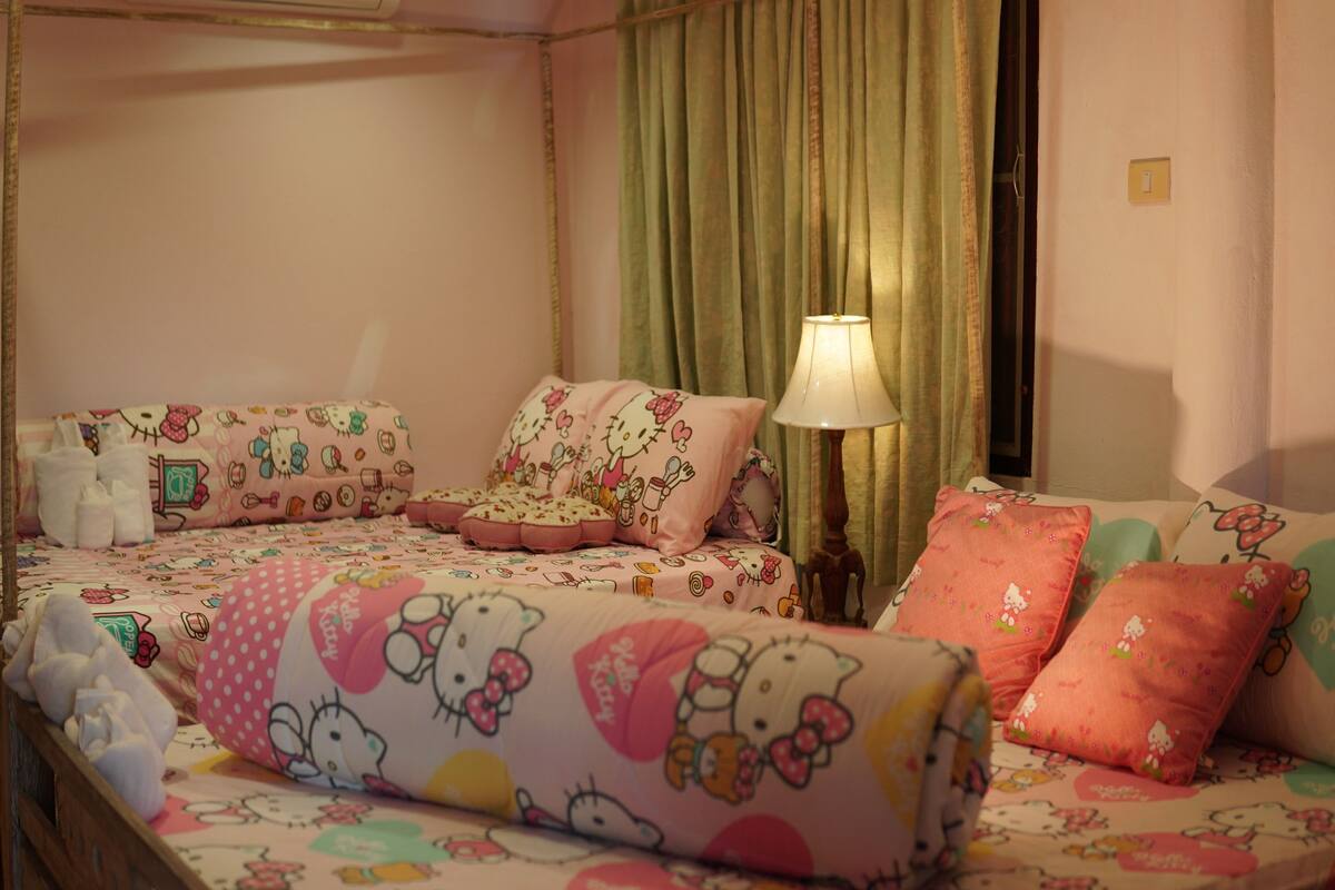 Una habitación con una cama con ropa de cama temática de Hello Kitty, una lámpara de noche y un cabecero de madera.