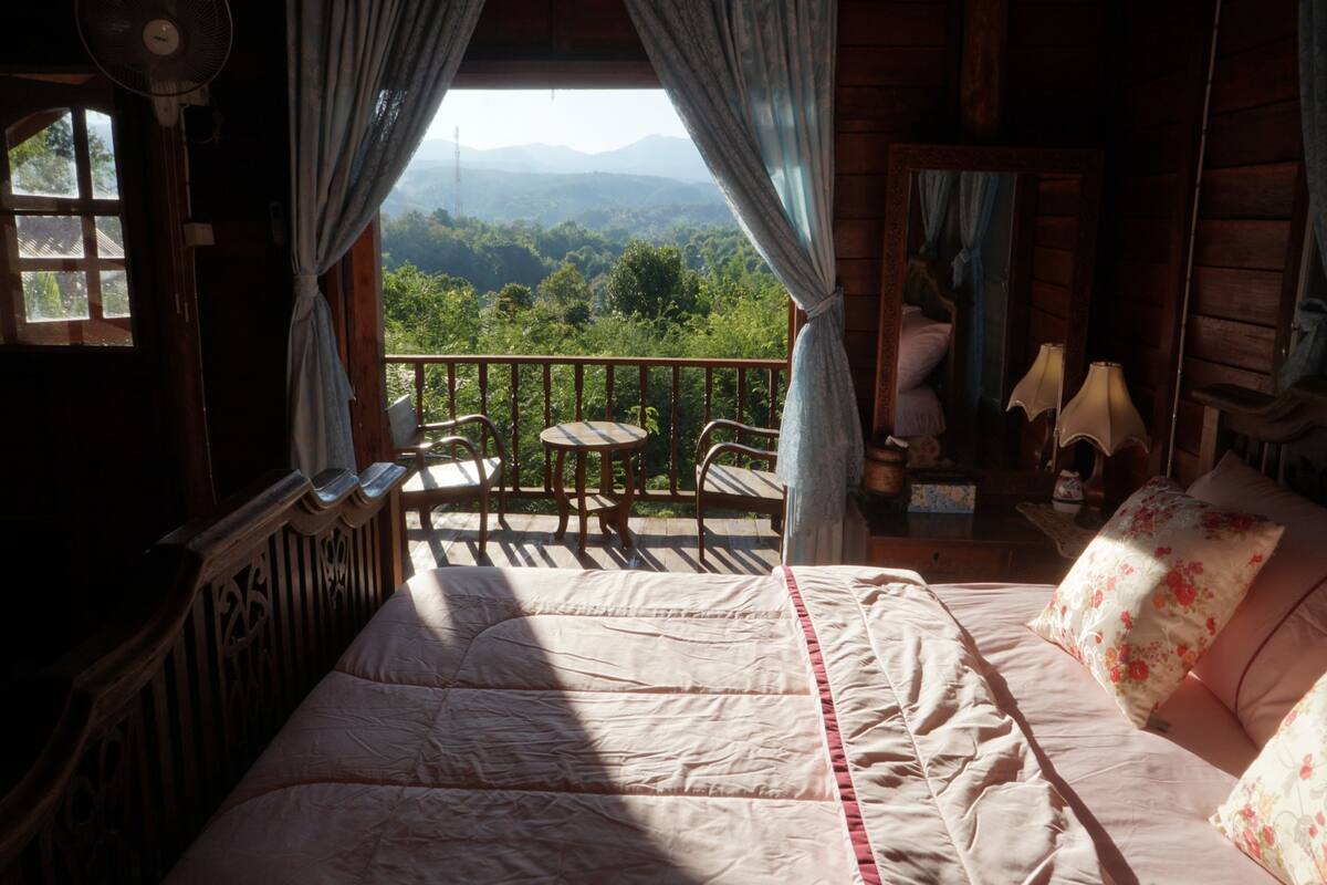 Una cabaña de madera con balcón, una cama con ropa de cama floral y vistas a una cadena montañosa boscosa.