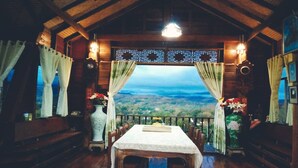 Un comedor rústico con una mesa lista para dos personas, bancos de madera y una vista panorámica a través de una ventana con cortinas.