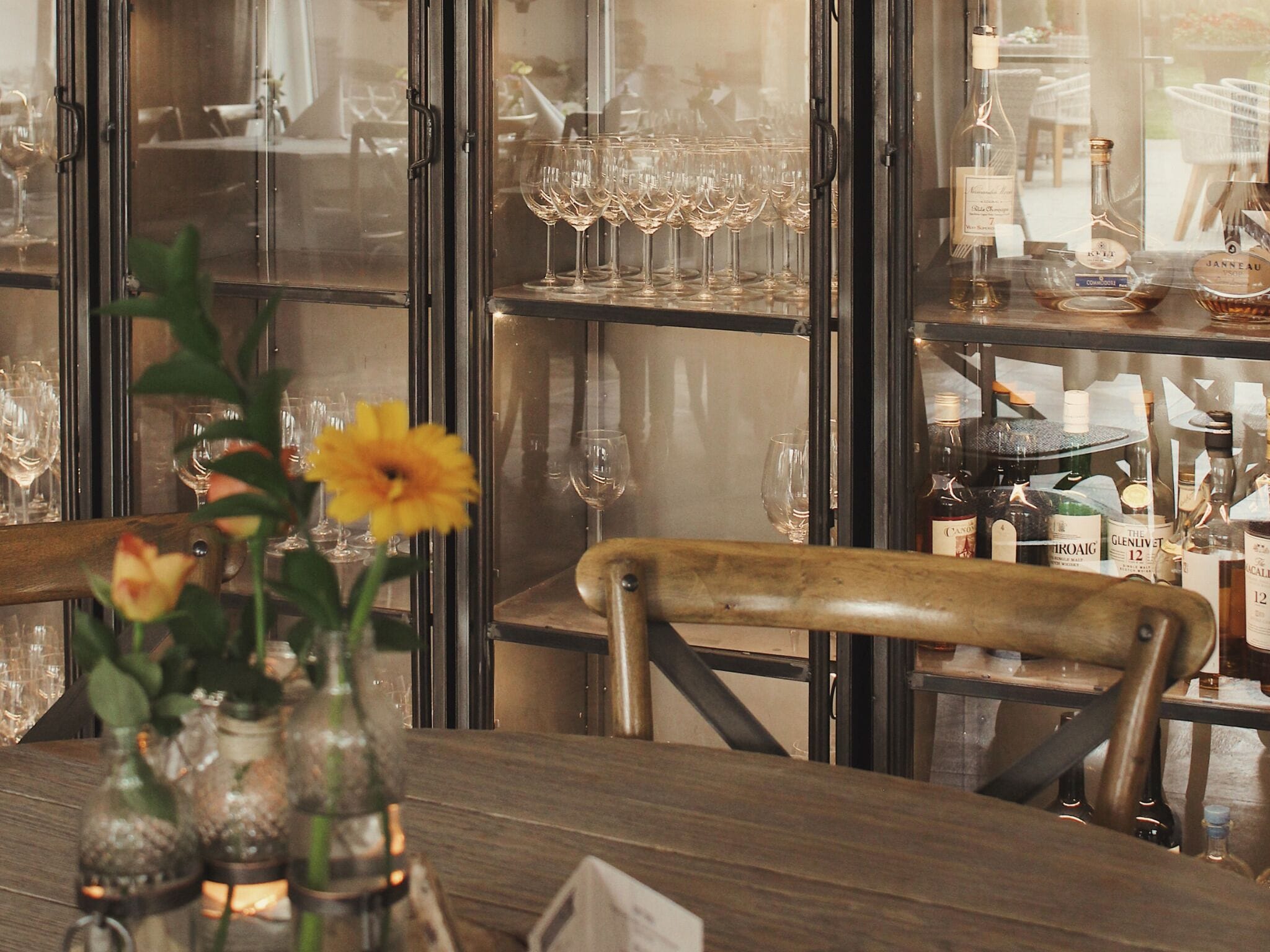 Una zona de bar con estantes llenos de copas y botellas de vino, una silla de madera y un jarrón con flores.