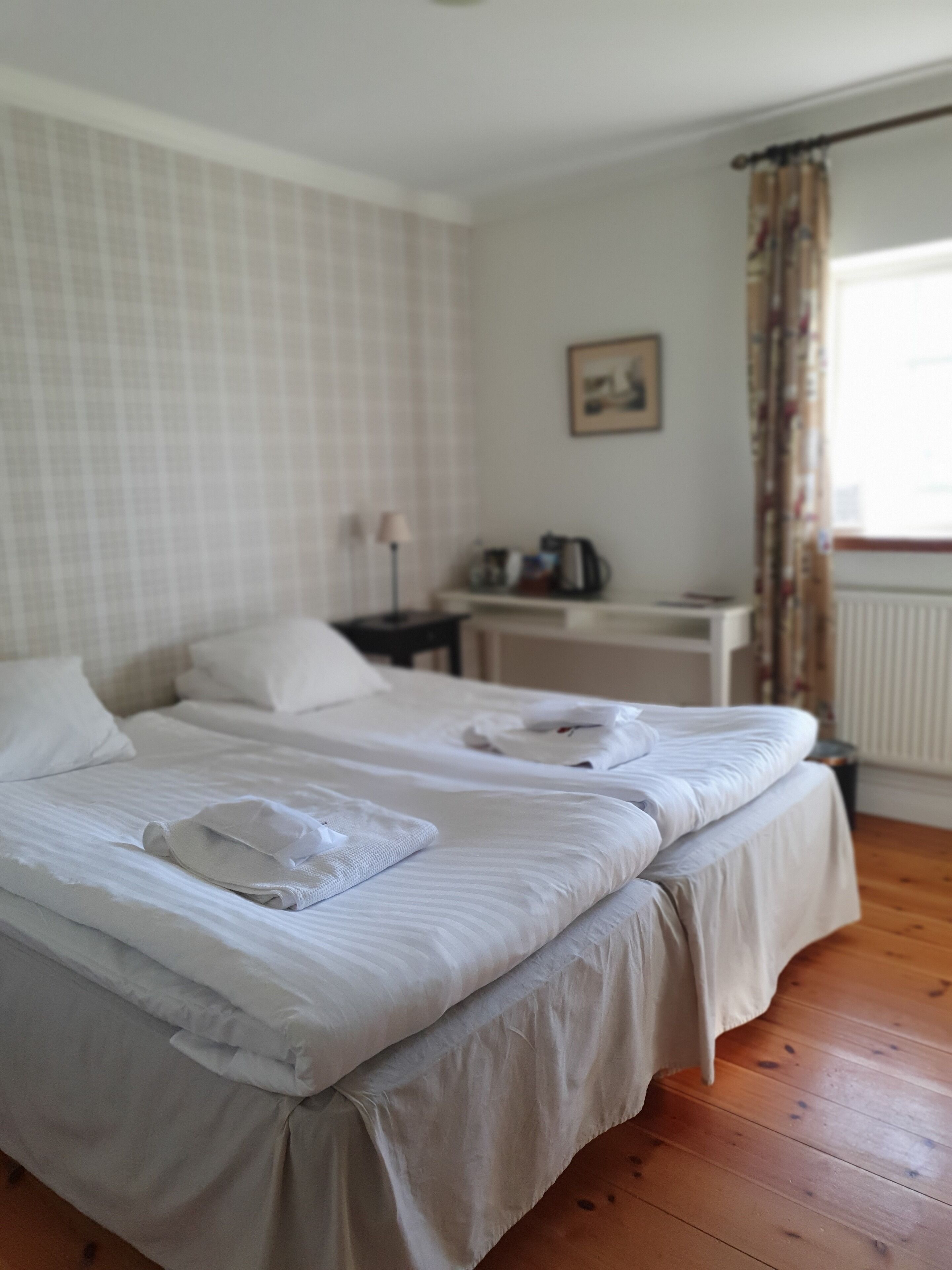 Habitación de hotel con dos camas, una mesita de noche con lámpara, una pequeña mesa con hervidor y una ventana con cortinas.