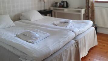 Habitación de hotel con dos camas, una mesita de noche con lámpara, una pequeña mesa con hervidor y una ventana con cortinas.