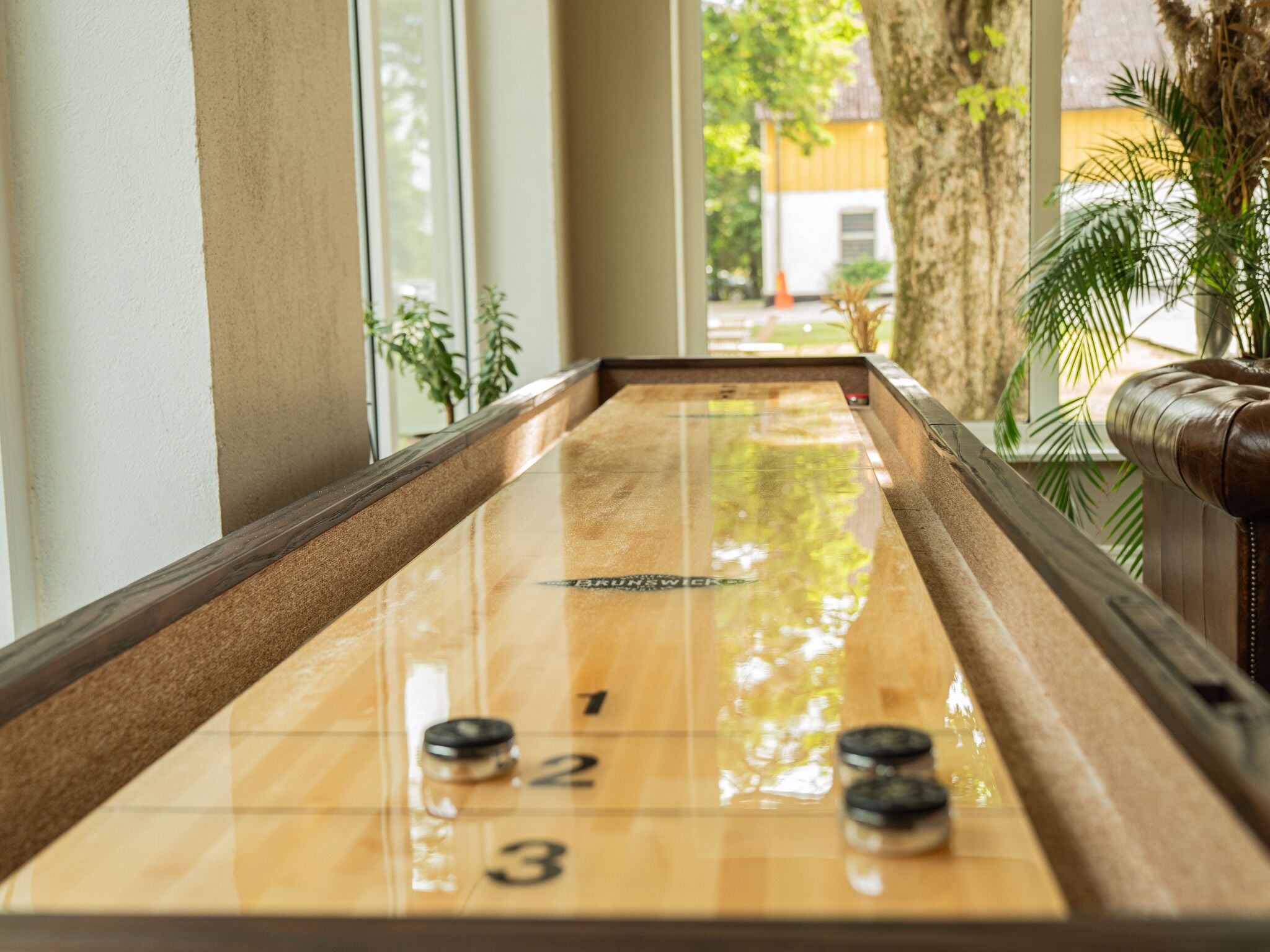 Una mesa de shuffleboard con áreas de puntuación numeradas, ubicada en un pasillo con ventanas amplias y plantas de interior.